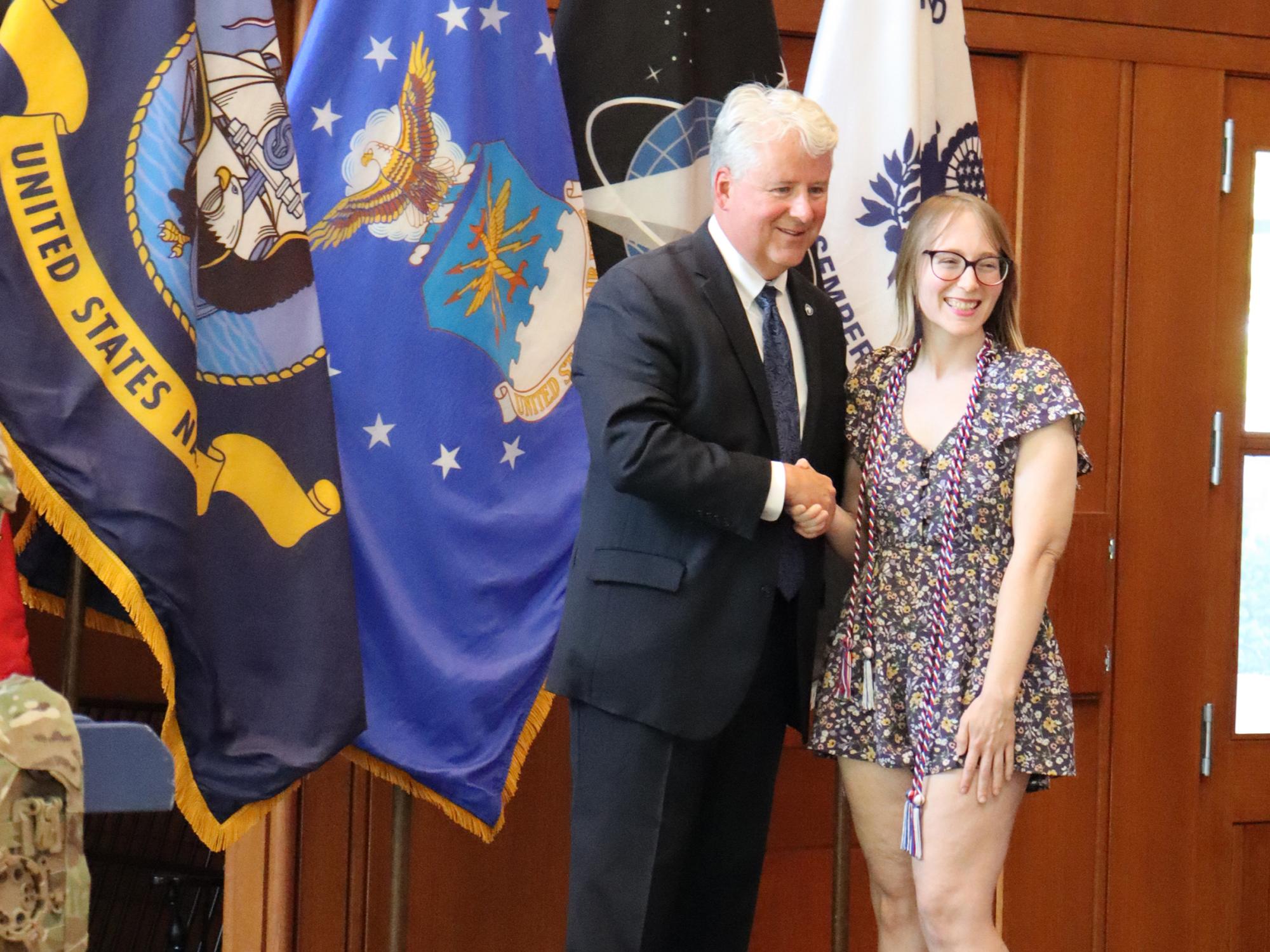 A woman shakes a man's hand in front of a row of U.S. military flags.