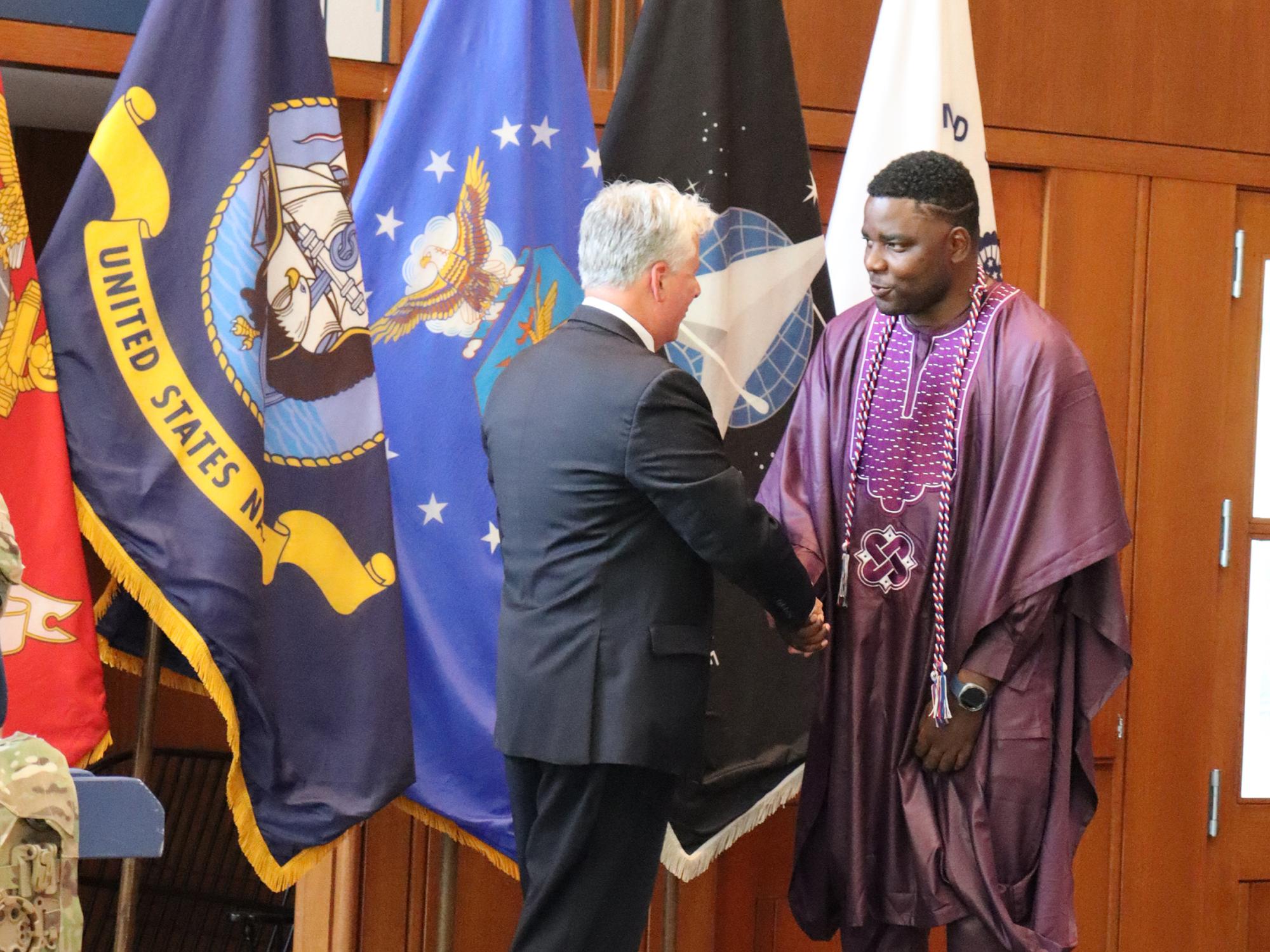 A man wearing purple cultural dress shakes another man's hand inside a room.