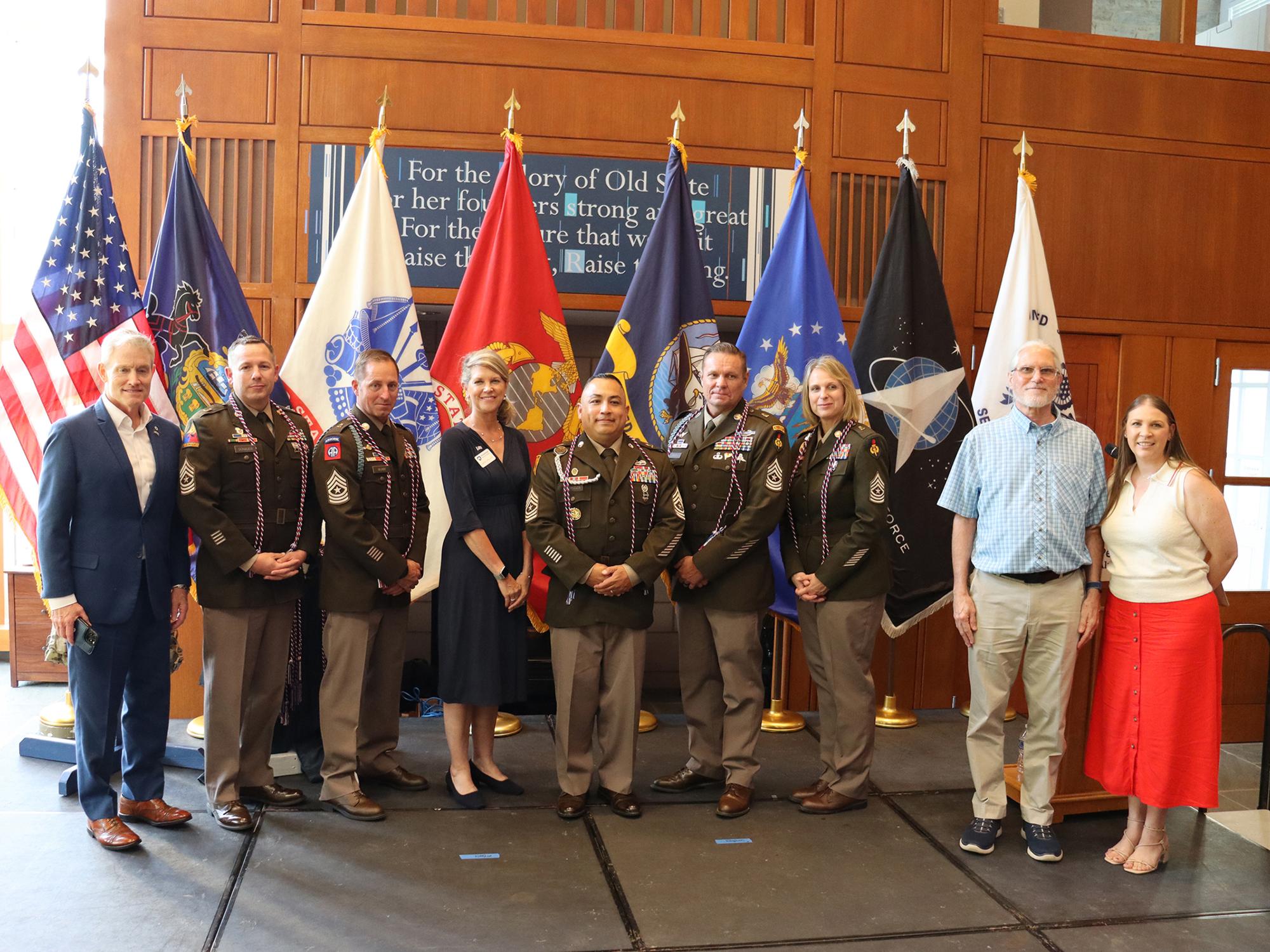 Nine people, some wearing military uniforms, pose in a room for a photo.
