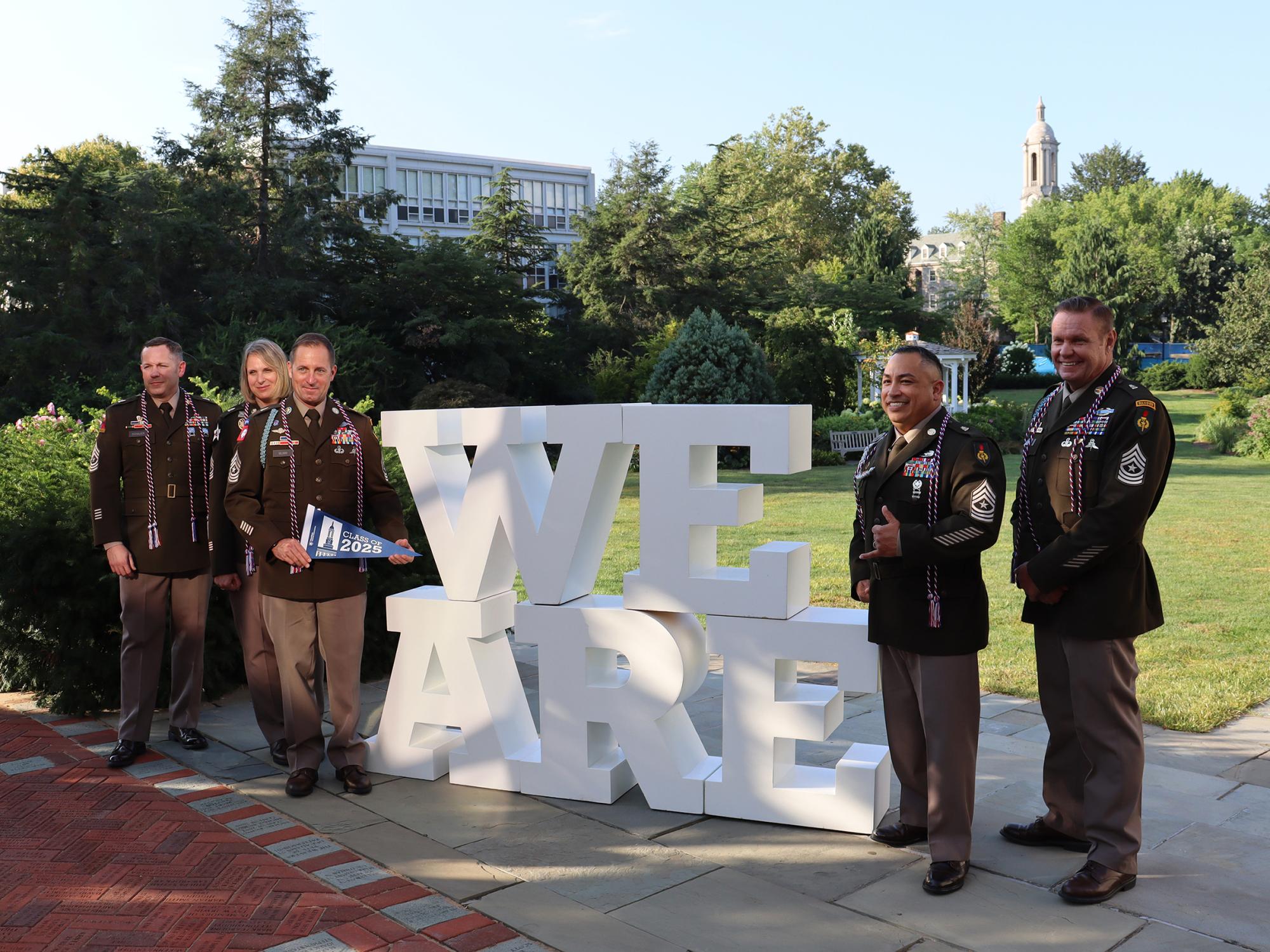 Five people wearing military uniforms pose in front of a "We Are" sign.