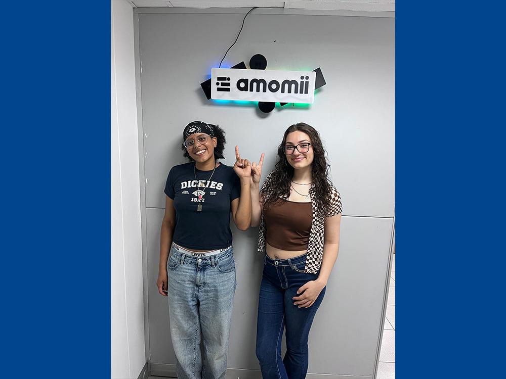 Two students standing on a white backdrop pointing towards a sign
