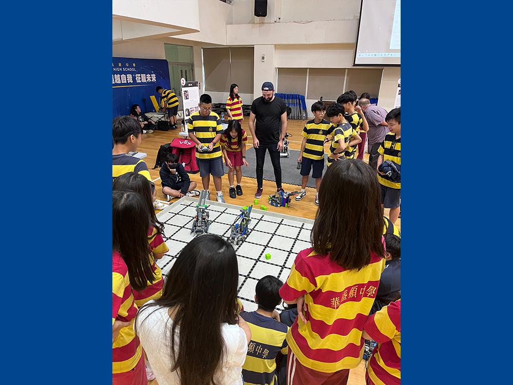 Person standing in a group of high schoolers looking at some robots in the center of a small pit.