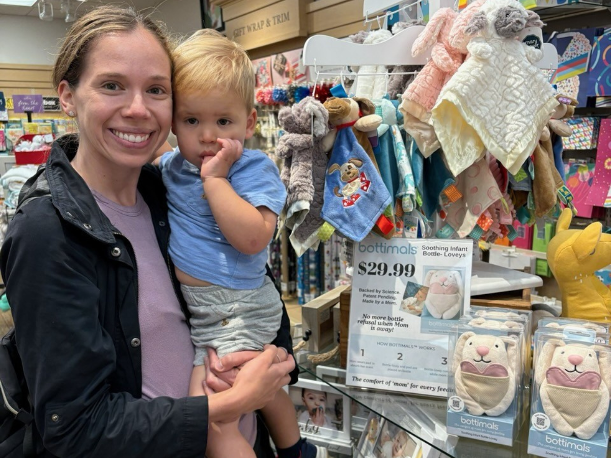 Julianne stands holding her toddler son in her arms in front of a shelf of baby products in a Hallmark store