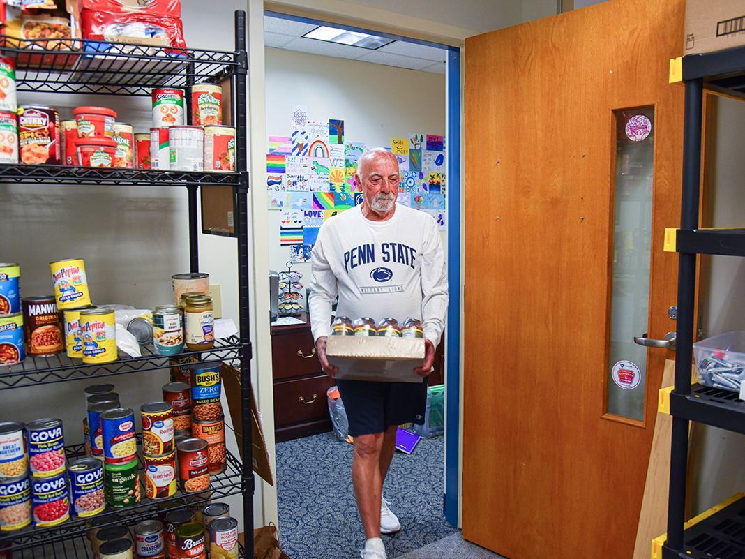 Man in a Penn State shirt carries a case of canned goods into a pantry stocked with food at Penn State Altoona’s Ivyside Eats.