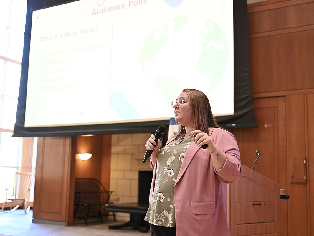 Ashlie Crosson, wearing a green shirt and pink jacket, stands in front of a projector screen while speaking into a microphone.