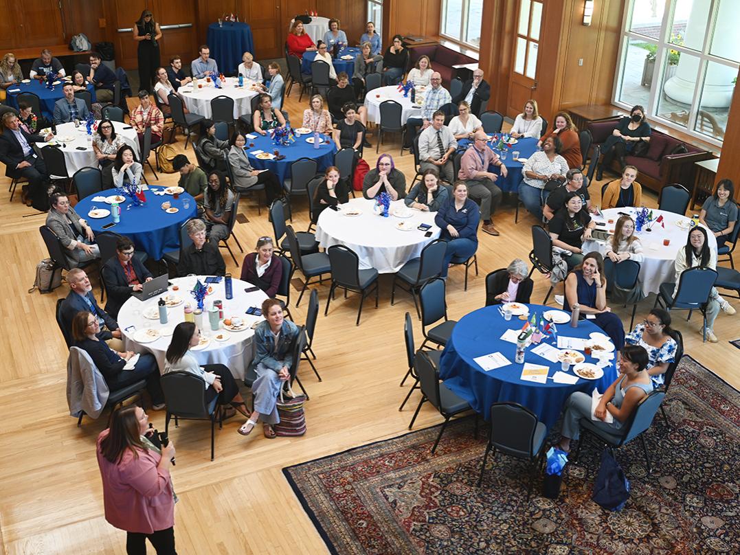 Small groups of people sit at round tables listening to a speaker.