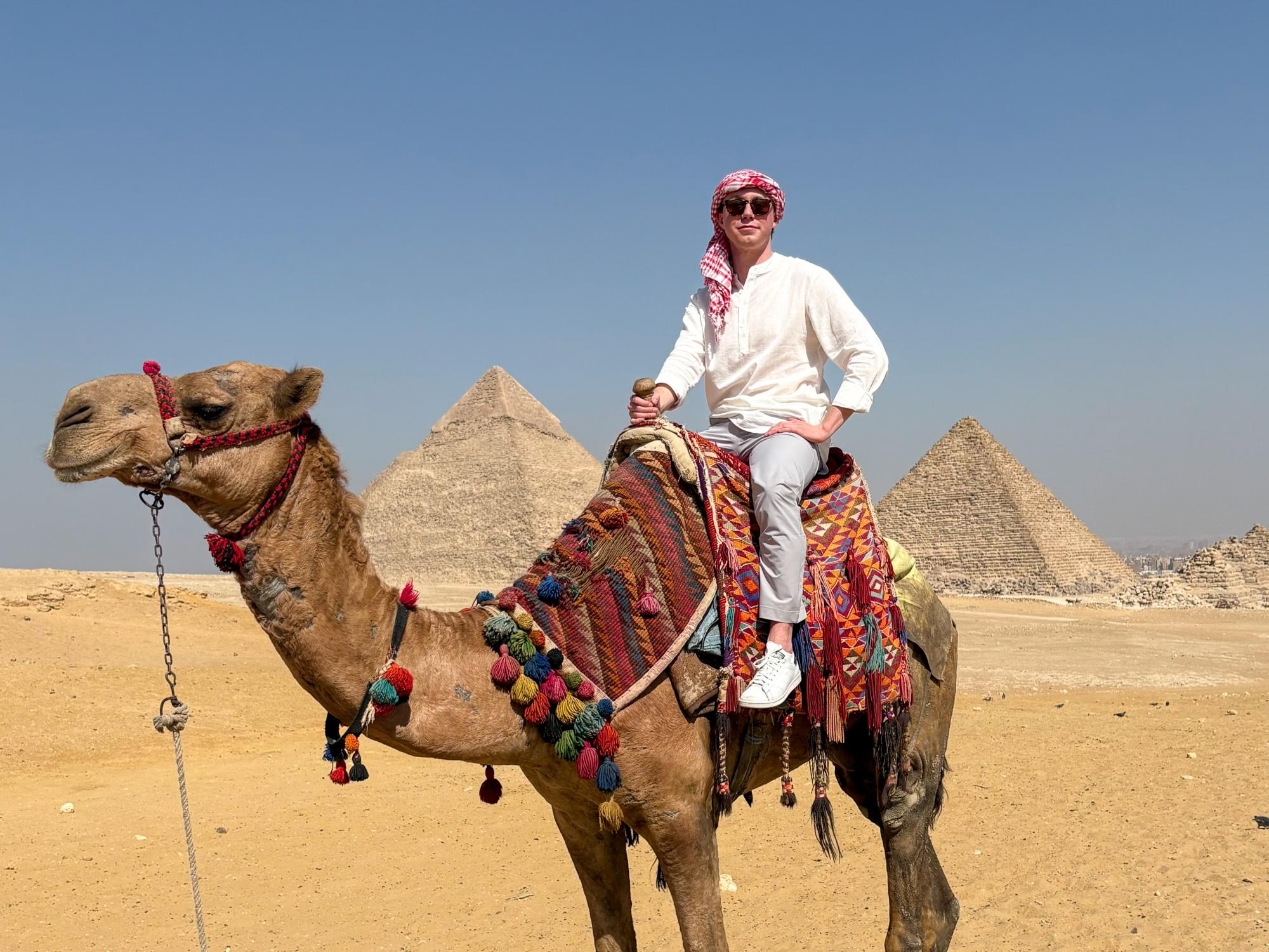 Zachary Graaf sits on a camel against a backdrop of pyramids in Egypt. 