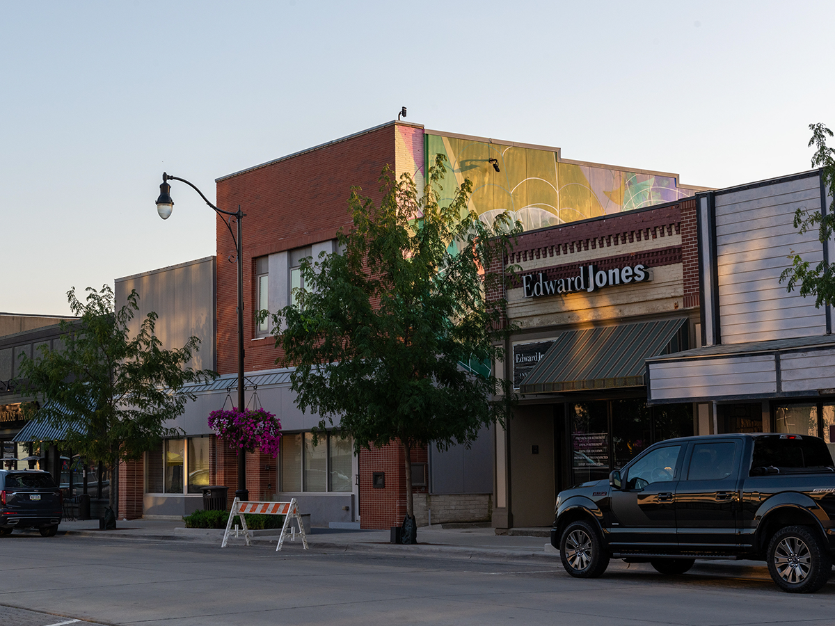 A view of downtown Indianola showing storefronts, trees, and a mural extending across the top of a red-brick building.
