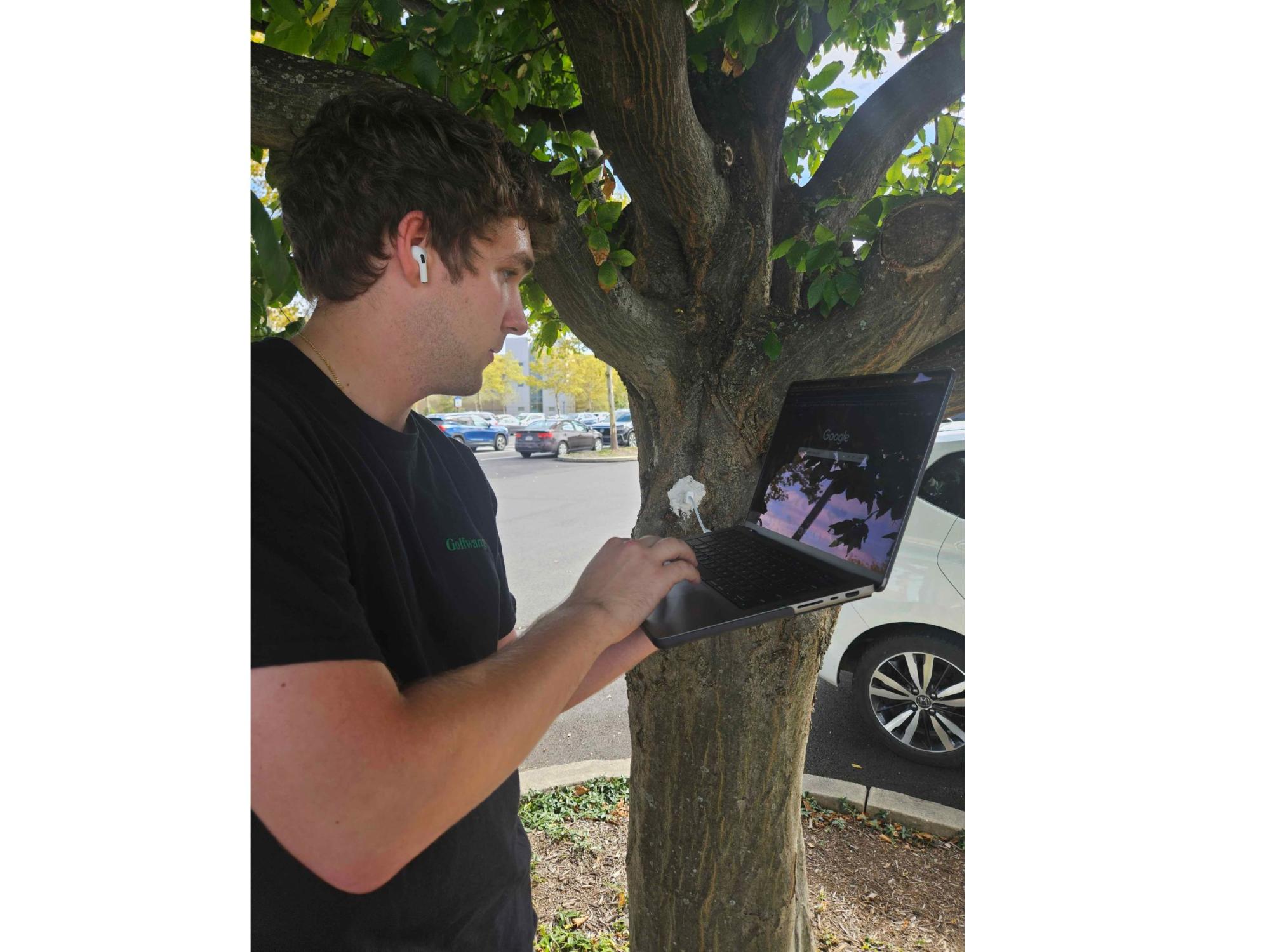 person in blue t-shirt and holding laptop stands next to a tree