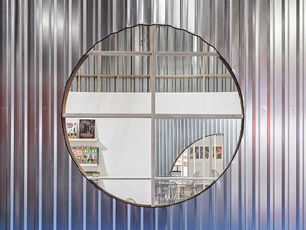 An aluminum ribbed wall with a circular mirror in the center that is reflecting the interior of a room.