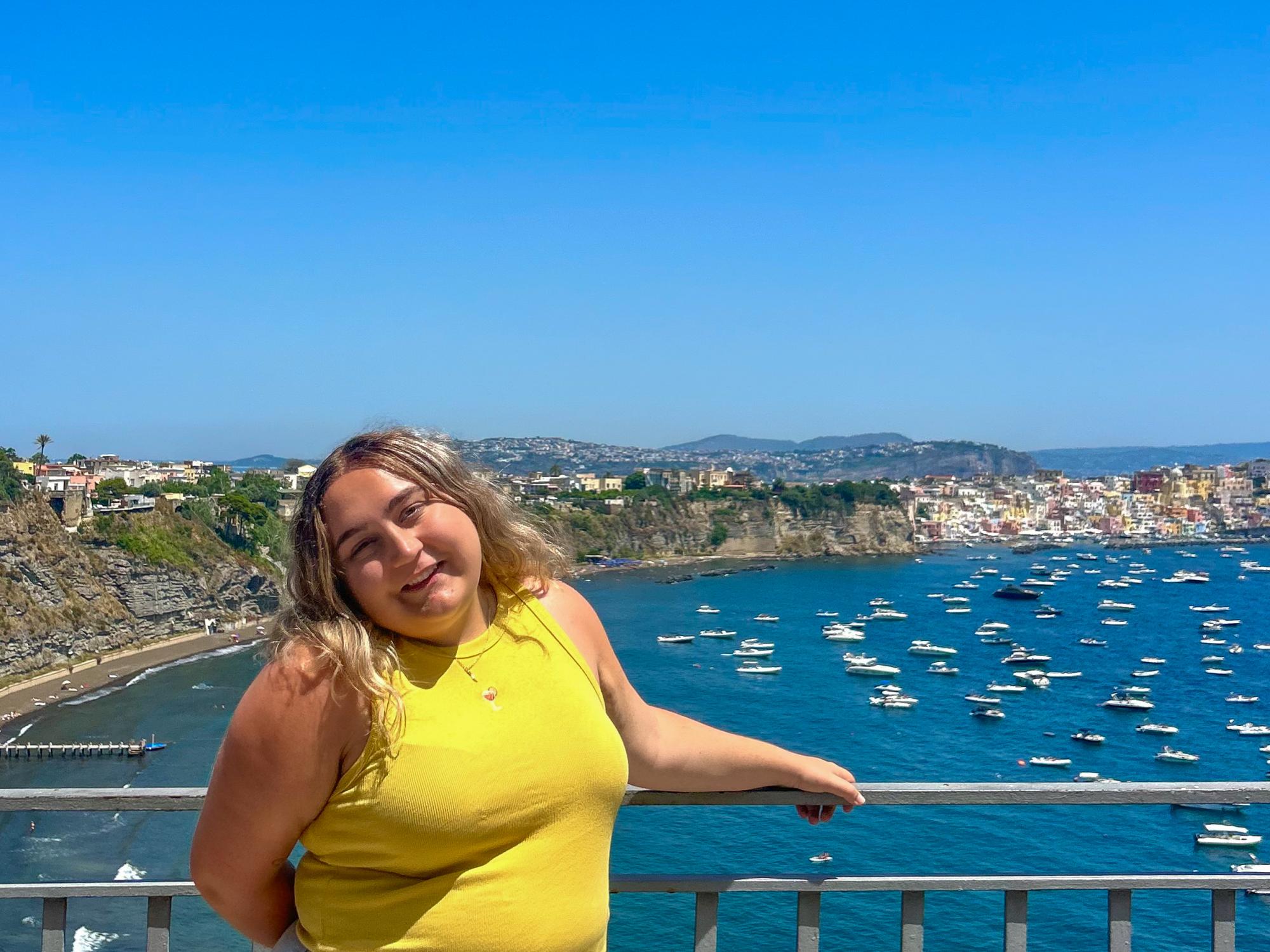 Katie Stum stands in front of a railing with the Procida Island coastline behind her.