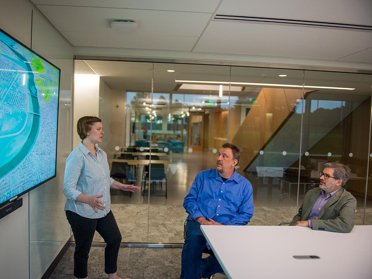 A woman standing at a screen with a colorful map speaks to two men sitting
