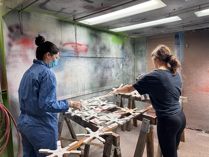 Two individuals spray painting wooden components in a shop. 