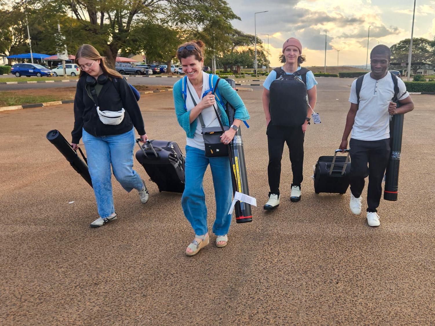 Four people carrying luggage walk with sunrise in the background.