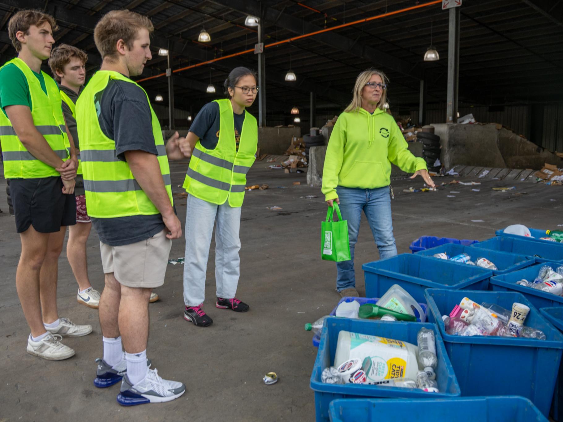 A group of people in safety vests stands in a recycling facility, observing bins filled with recyclables.