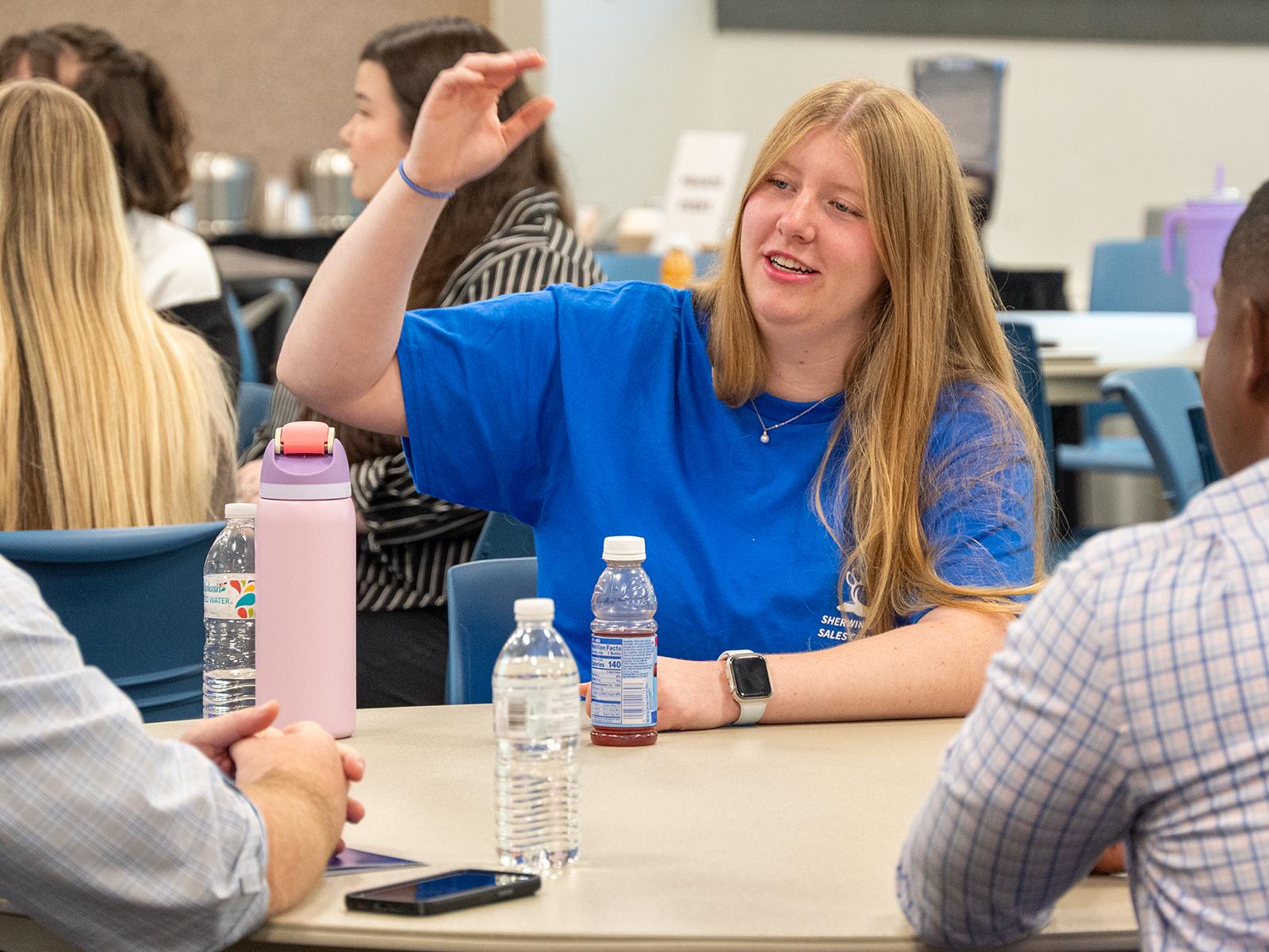 A student in a blue shirt gestures, while sitting at a table with others