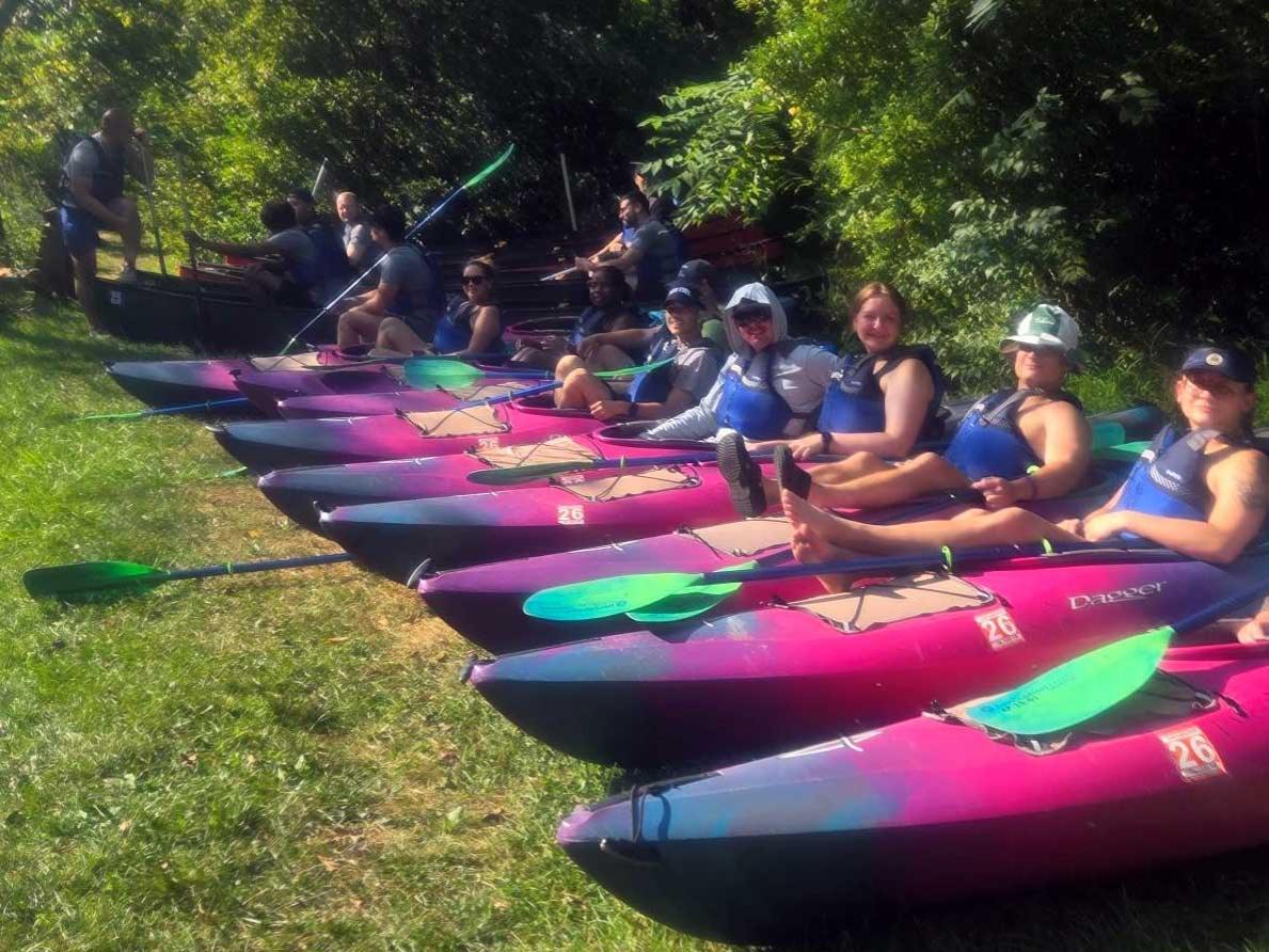 A row of people sitting in bright pink and blue kayaks on the grass