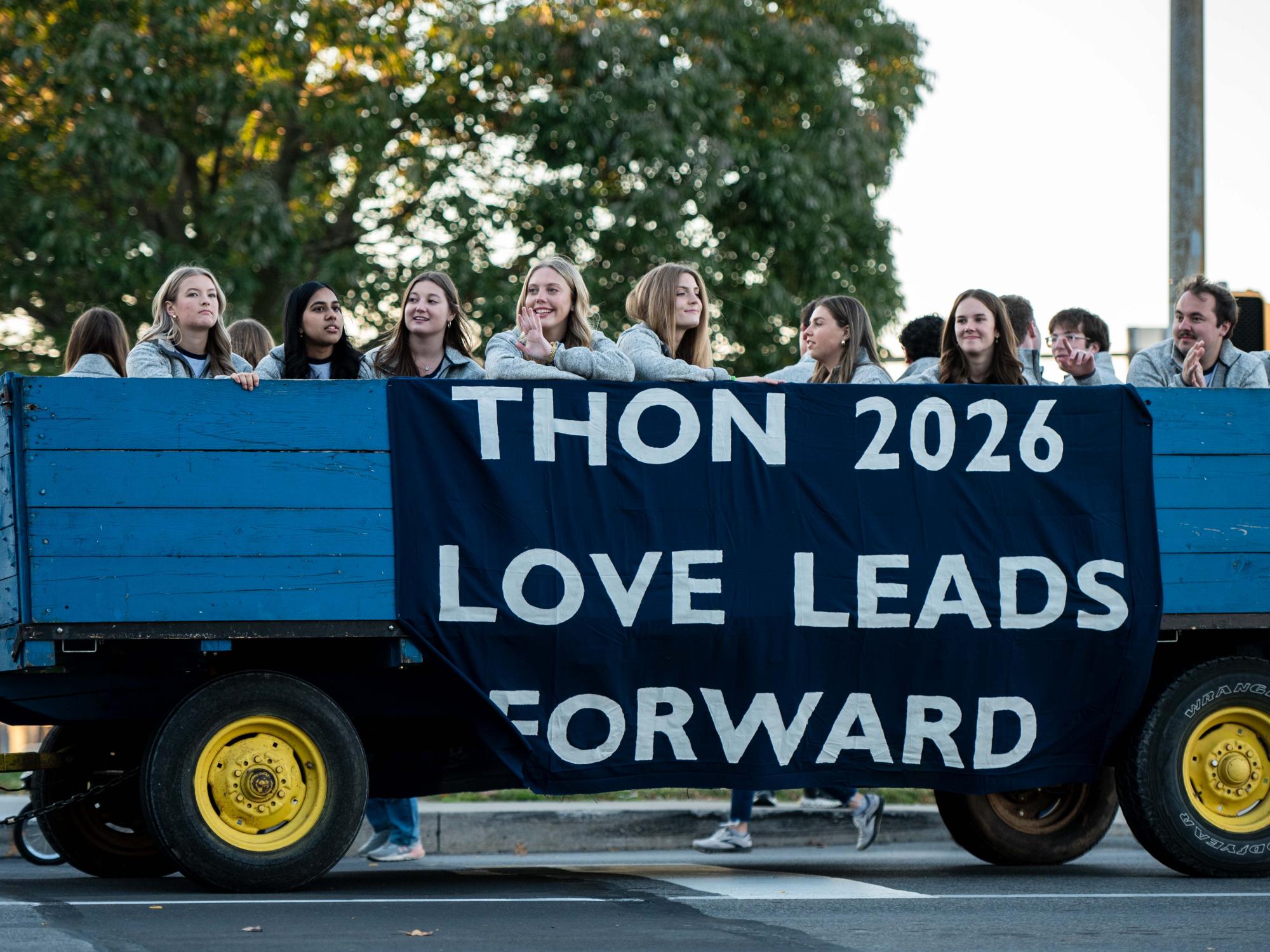 A group of people ride on a blue trailer with a large banner reading “THON 2026 Love Leads Forward” during a parade or event.