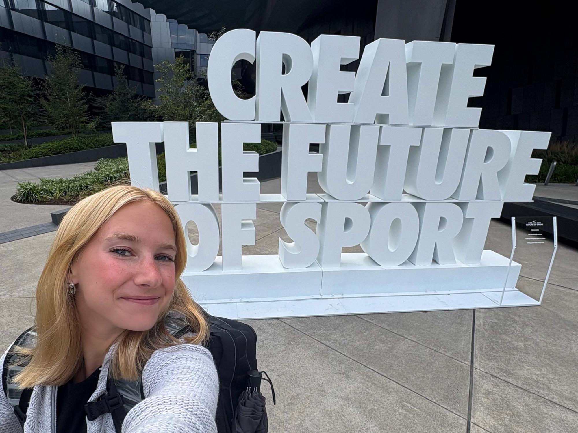 Katie Dallas stands outside the Serena Williams Building at Nike World Headquarters in Beaverton, Oregon, with a sign behind her that reads “Create the future of sport.”