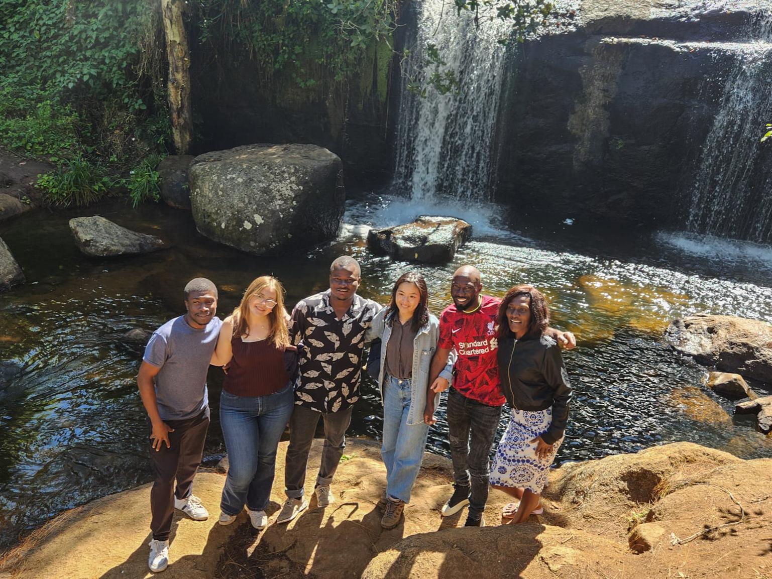 A group of people stands together in front of a cascading waterfall, surrounded by lush greenery and mist.