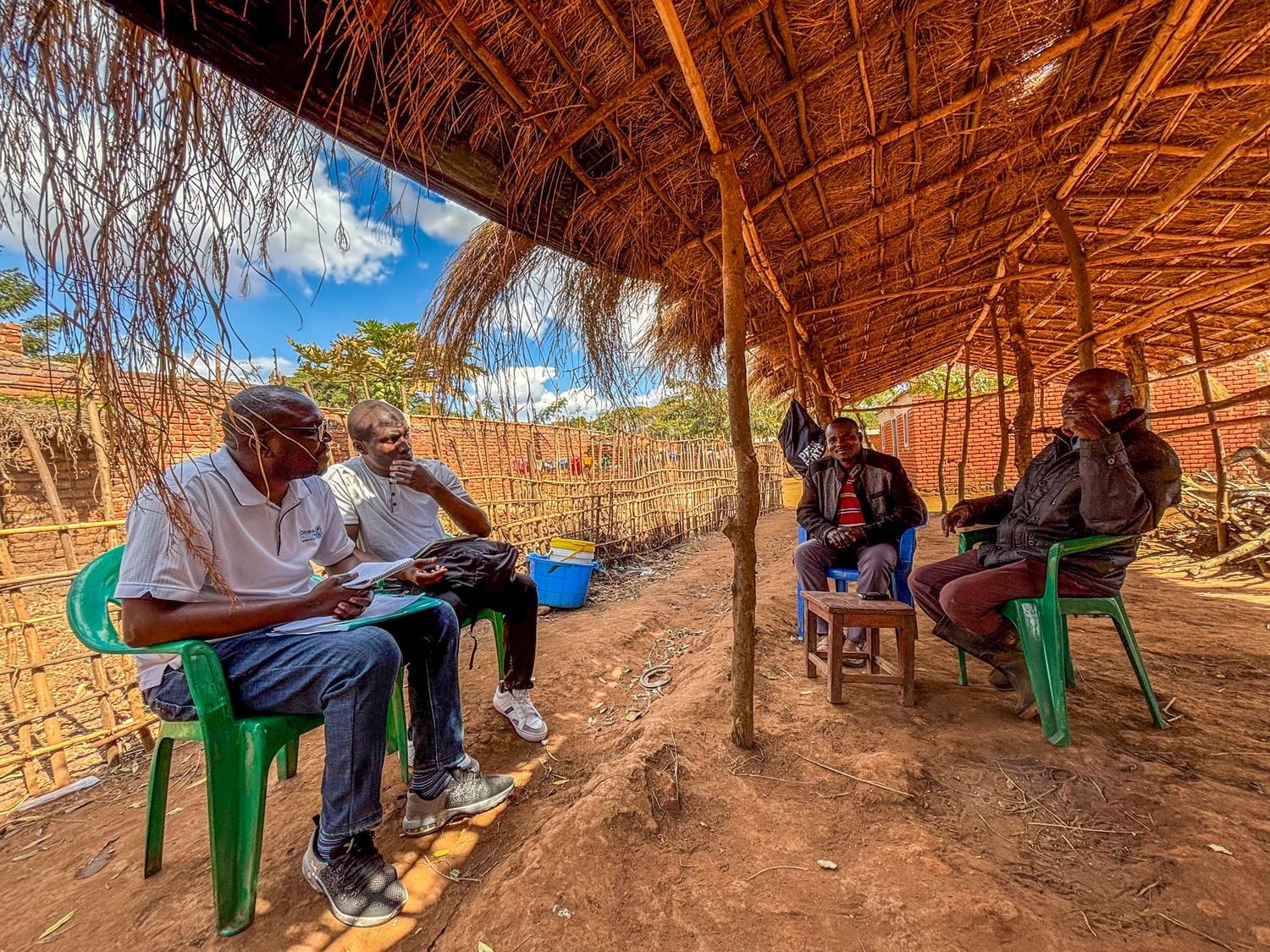 Four men engage in conversation under a thatched-roof shelter with wooden supports. They sit on green chairs and a stool on a dirt floor.
