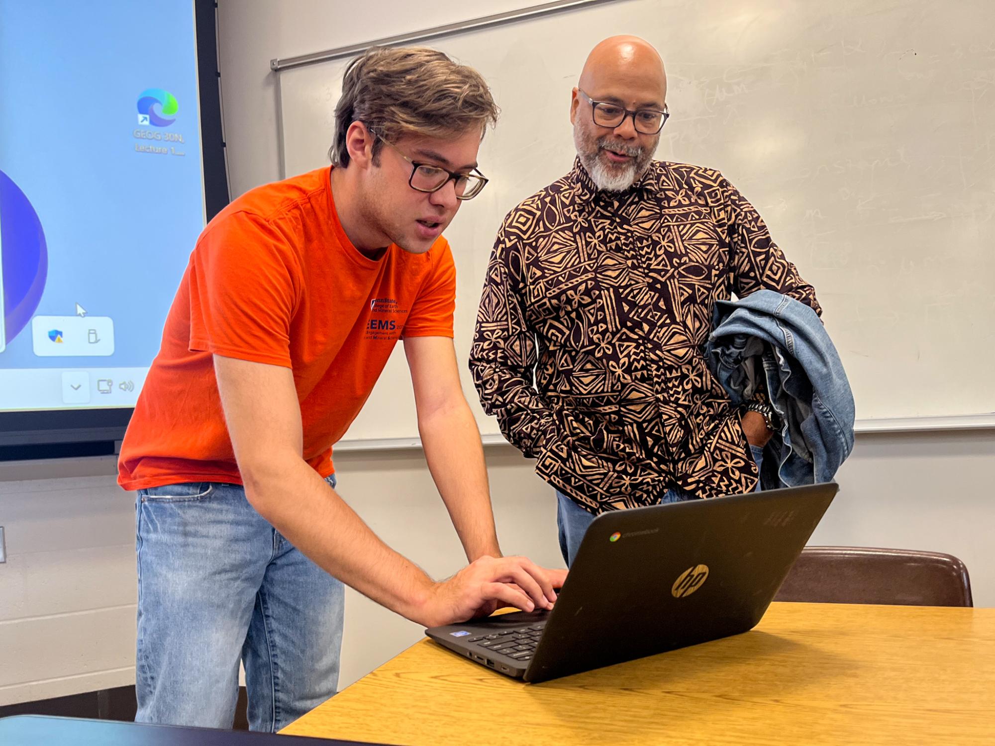 A young man in an orange T-shirt is focused on a laptop, while an older man in a patterned shirt observes.