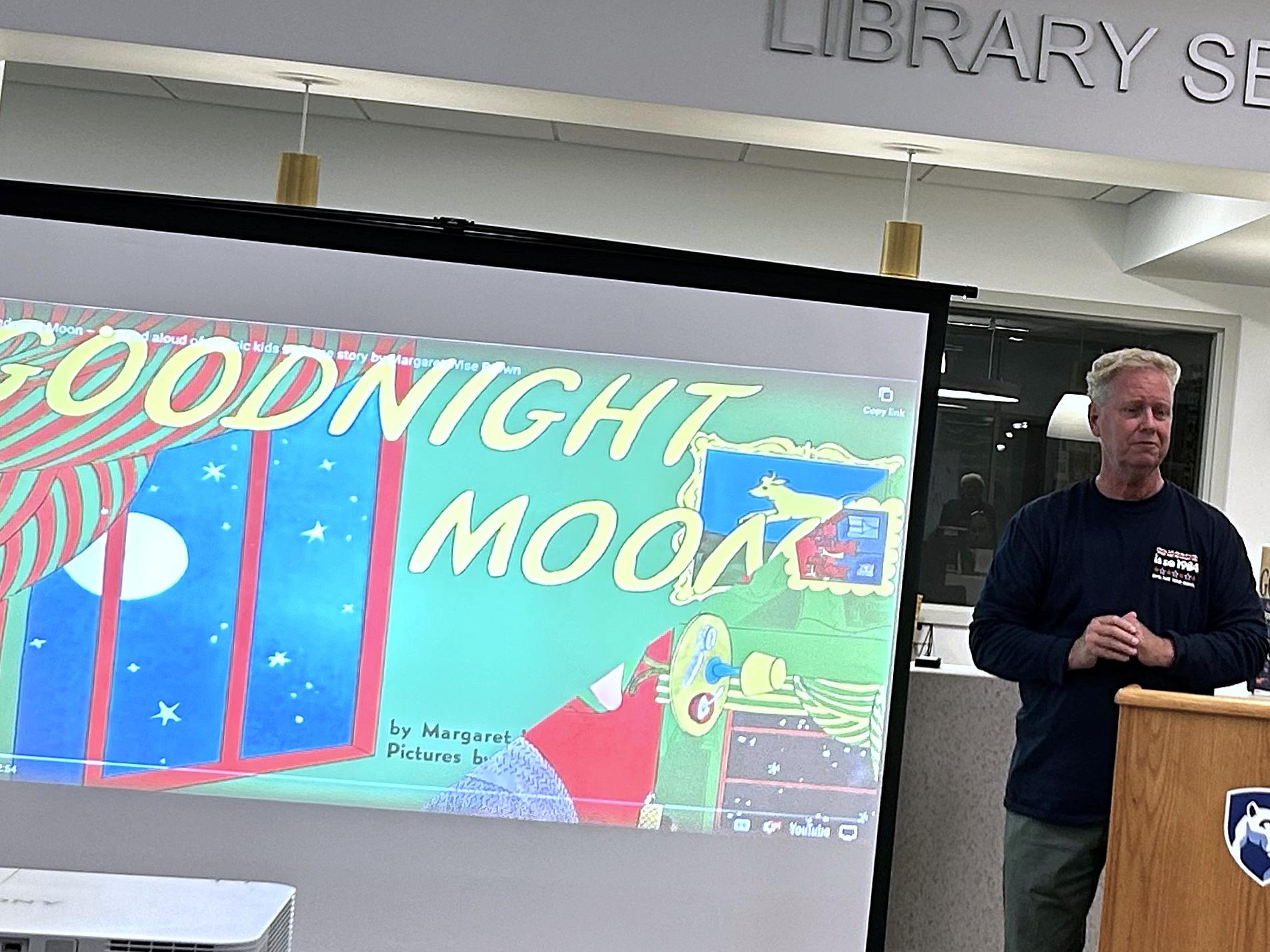 man stands in front of projection screen with the cover image of the book Goodnight Moon 