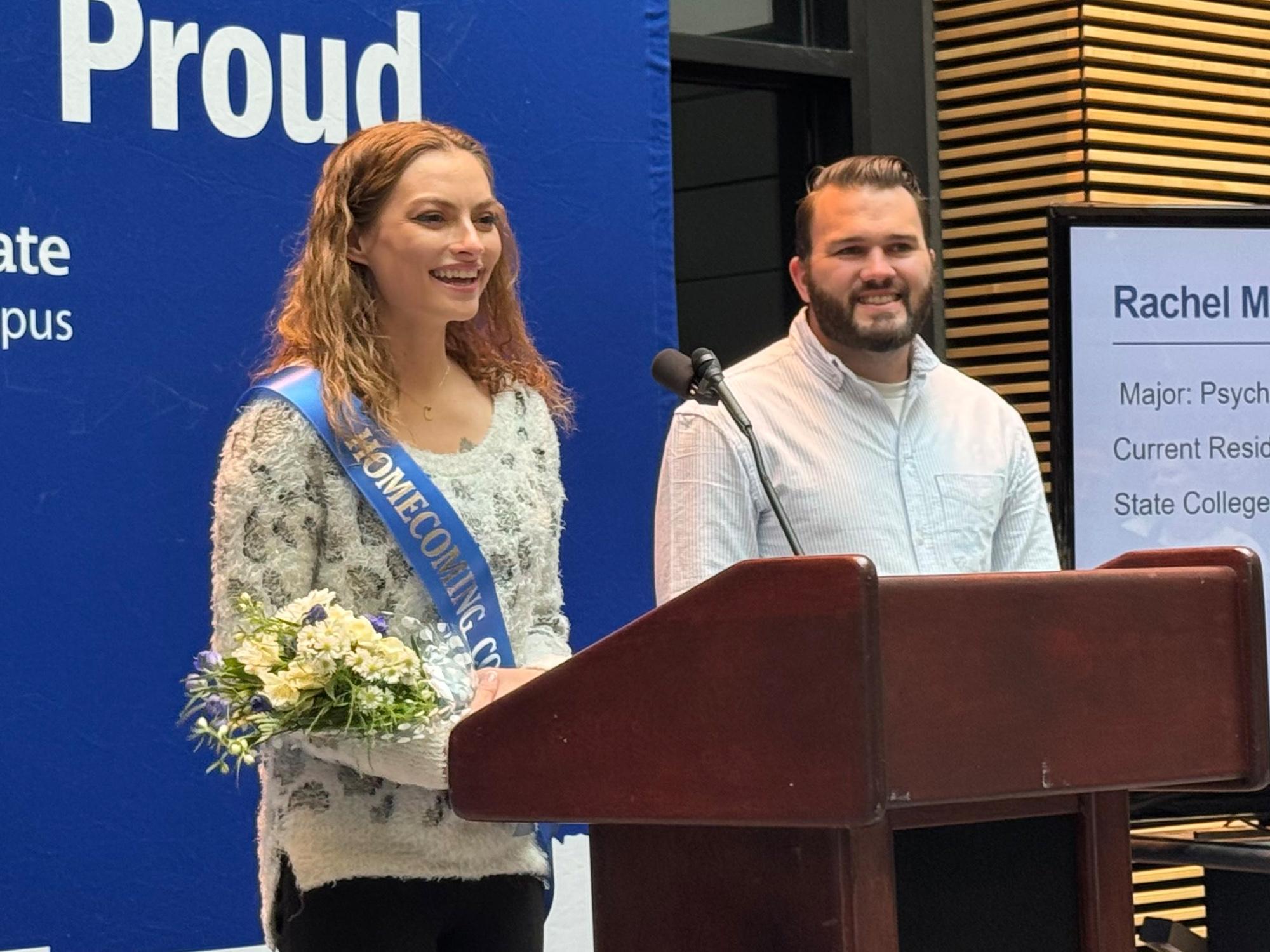 A woman wearing a Homecoming Court sash stands at a podium, holding a bouquet of flowers.