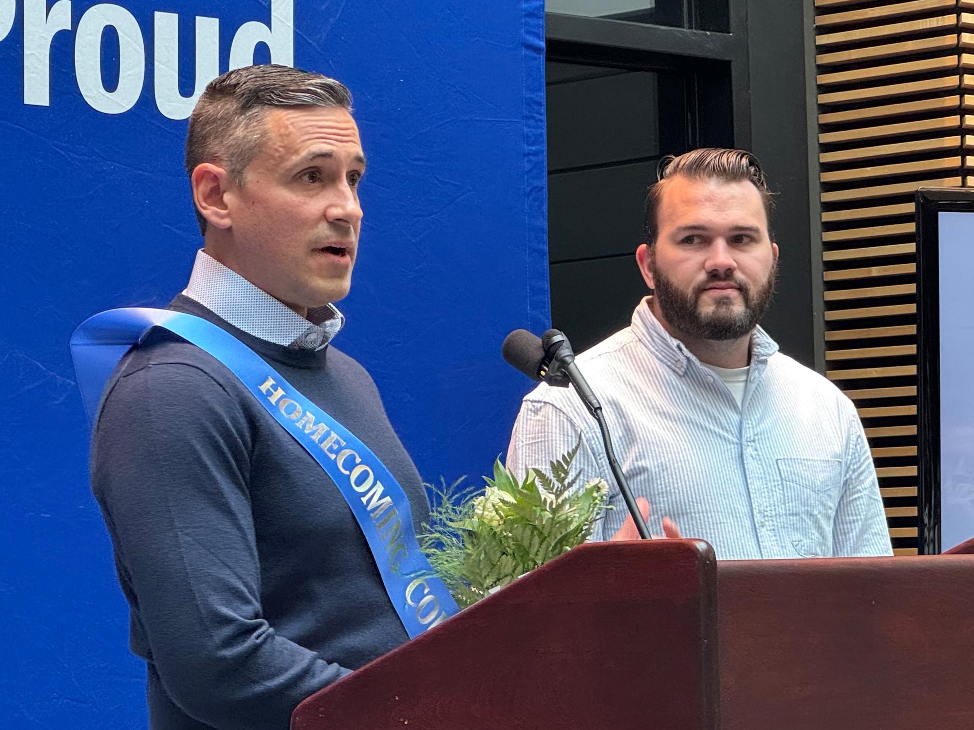 A man wearing a homecoming sash stands at a podium