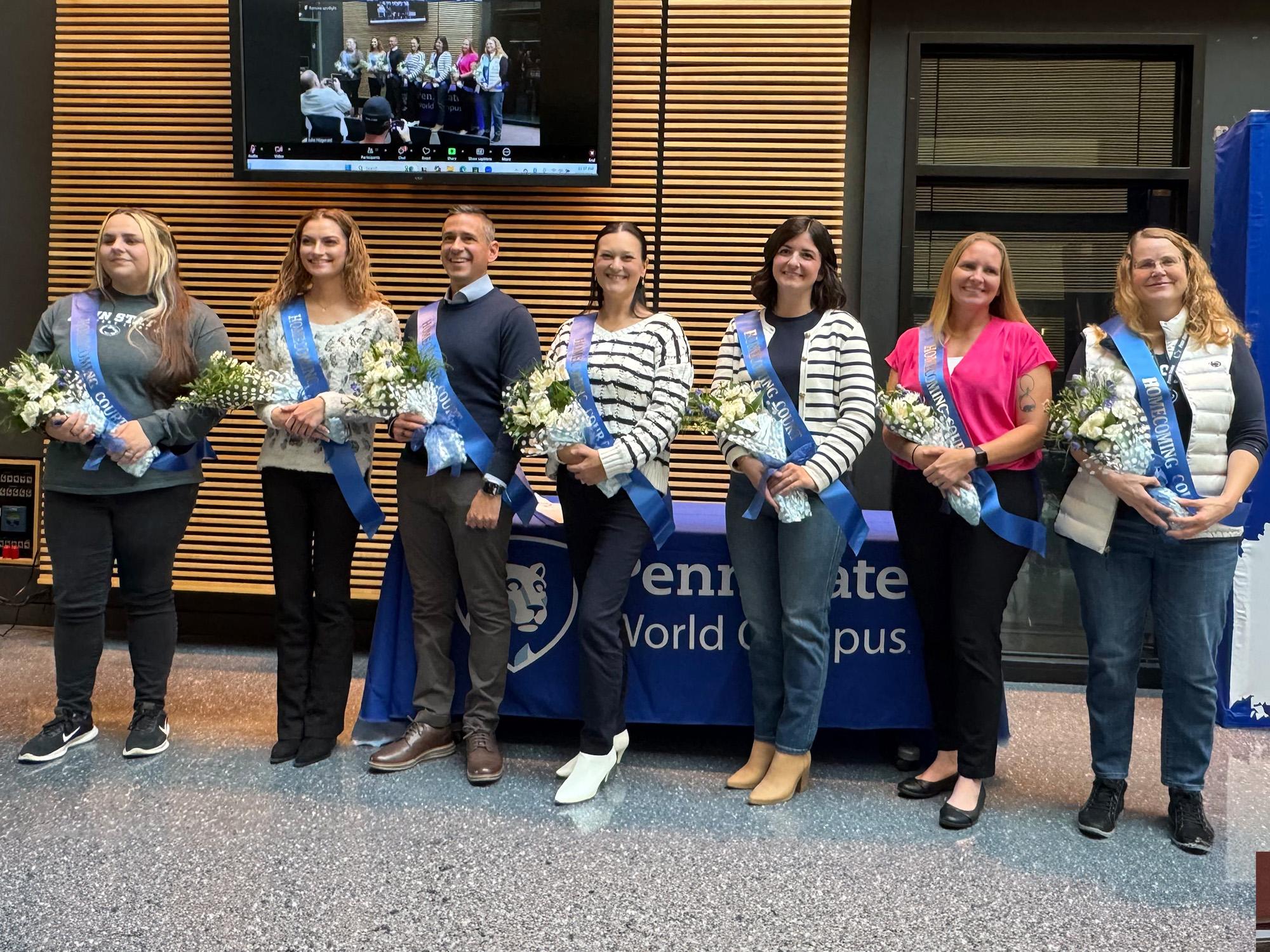 Seven people wearing sashes and holding bouquets stand in front of a table.