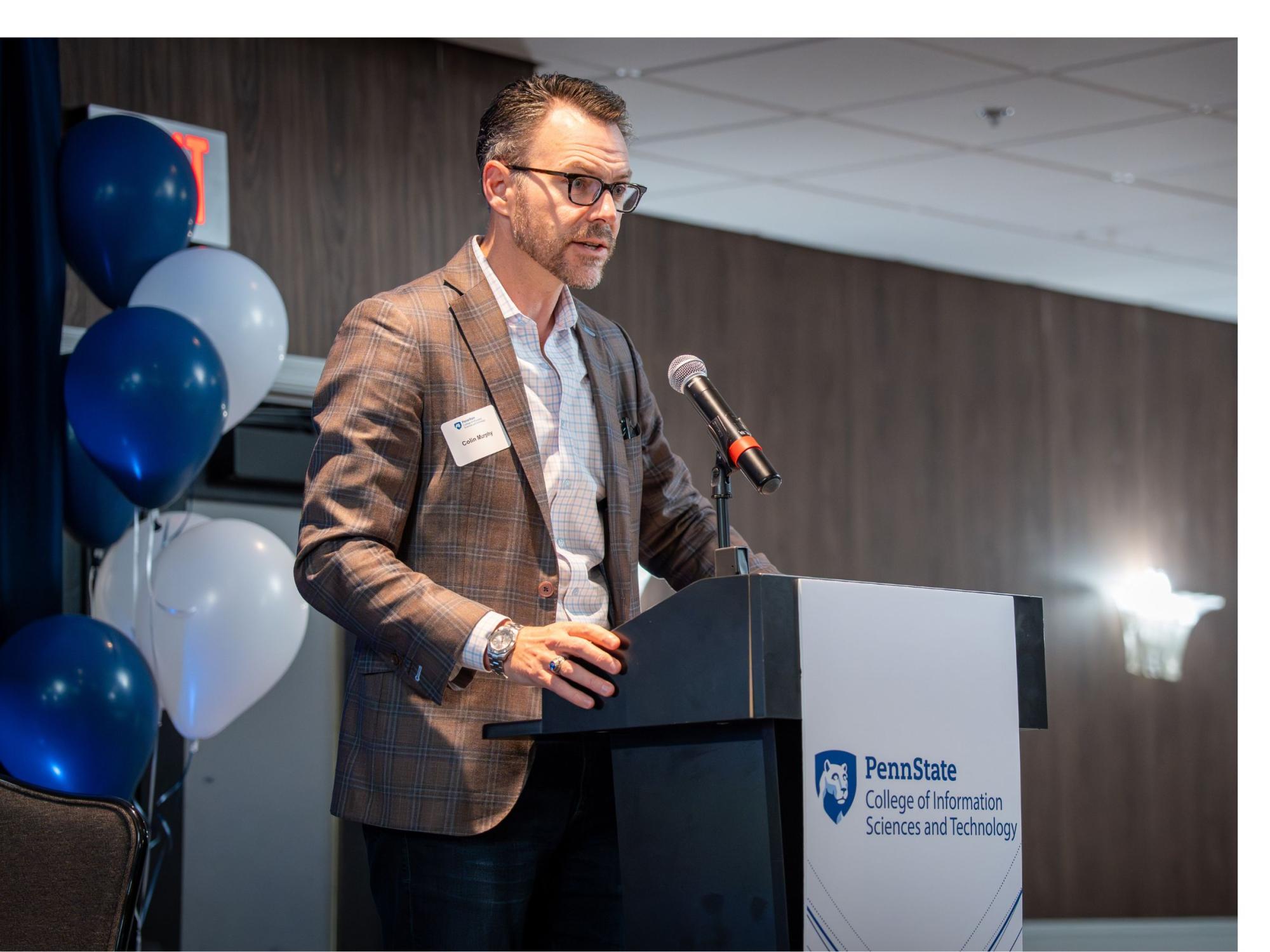 person with short hair and glasses stands at podium with balloons in background