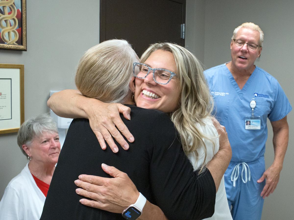 Dr. Marnie Kaplan smiles as she hugs a woman patient, who is seen from behind. Dr. Chance Kaplan, standing on the right, and a woman, seated on the left, look on. Dr. Marnie Kaplan has long hair and is wearing glasses and a top. The woman patient is wearing a sweater. Dr. Chance Kaplan is wearing scrubs and has a name badge on a lanyard attached to his pocket. 