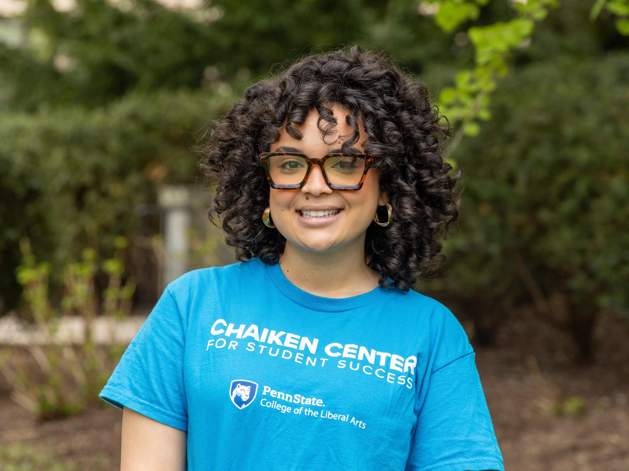 Alexa Sanchez poses outdoors for her peer success coach headshot wearing a sky blue shirt with Chaiken Center for Student Success and the Penn State College of the Liberal Arts logo.
