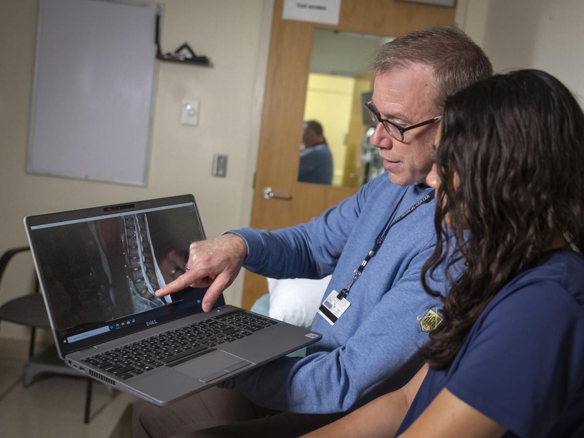 Dr. Matthew Silvis points to an X-ray image of a spine on a laptop while a teen girl next to him looks at it. He is wearing a long-sleeved shirt and a nametag with a lanyard. The girl has long hair and is wearing a T-shirt.