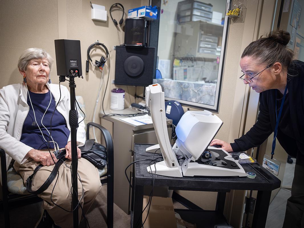 A woman sits in a chair on the left with cords hanging from hearing aids in her ears. Dr. Jackie Price, right, leans over a computer. Behind them are speakers, headphones and other audiology equipment.