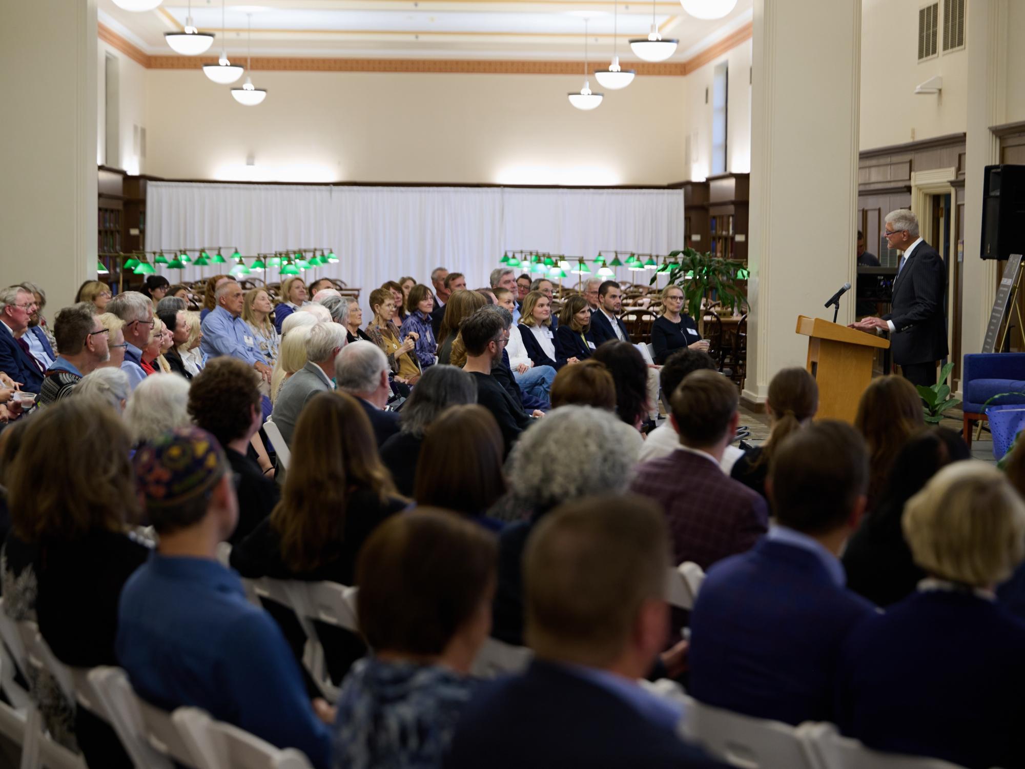man speaks to a large seated crowd in a large room