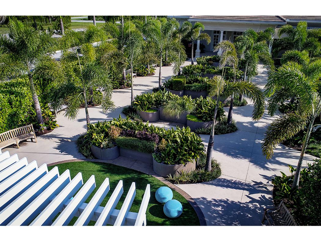 Aerial view of a landscaped park with neatly arranged palm trees, planters, benches, and walking paths, adjacent to a modern building.