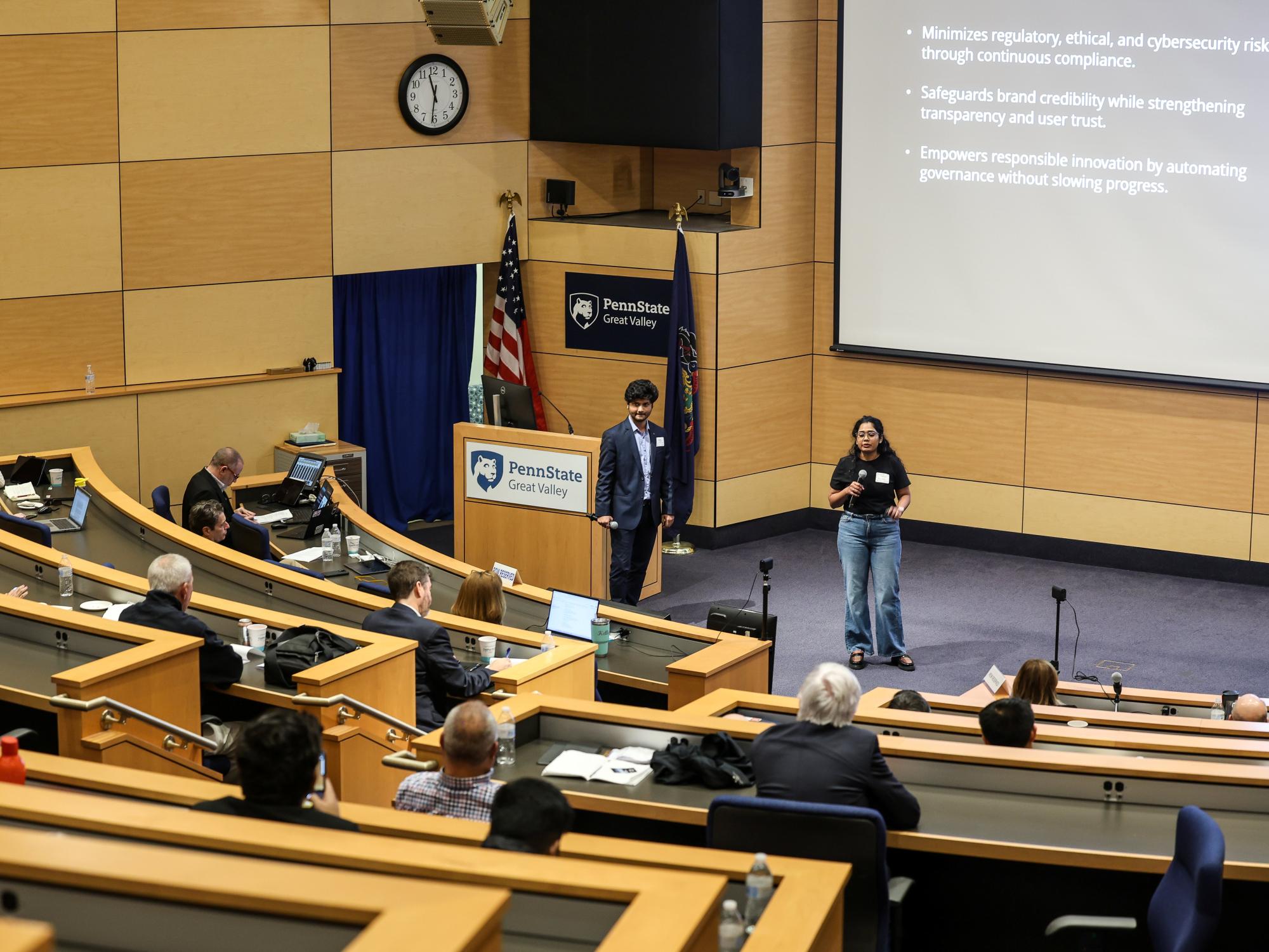 Two people present on a stage in front of a slideshow to a group of people in an auditorium.