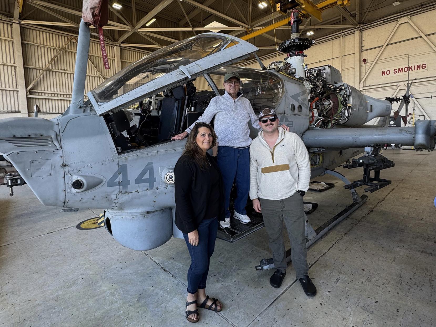 Three people stand next to a military helicopter in a hangar