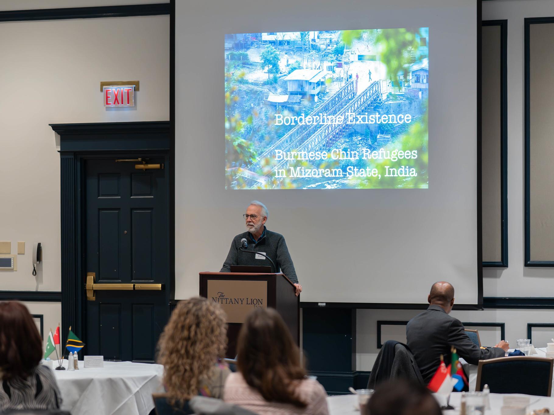 A man at a podium presents a slideshow at an event for an audience