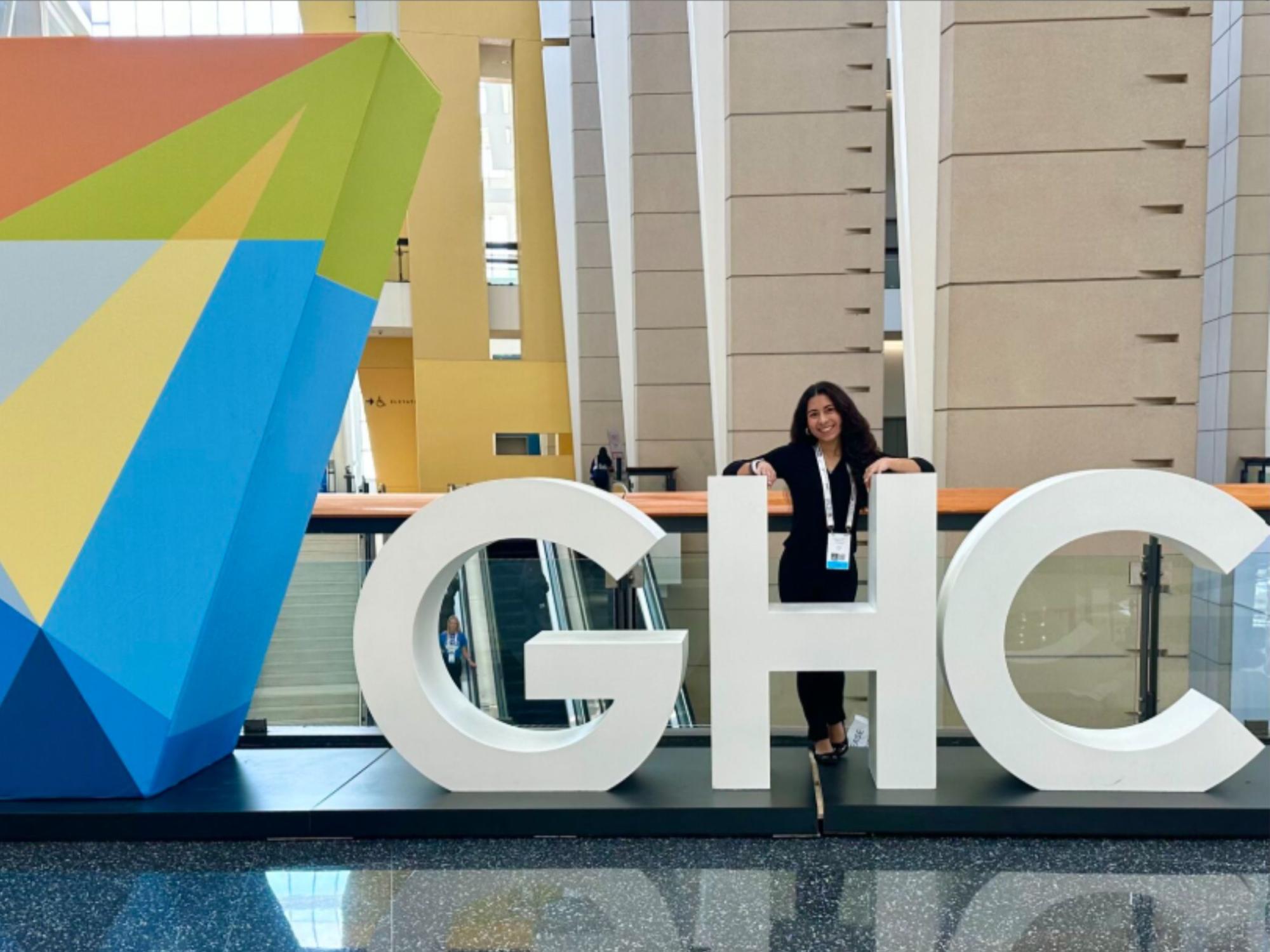 smiling person with long dark hair stands next to large letters at conference center