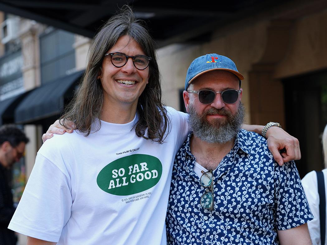 Man in white T-shirt with green logo on chest and his left arm around a man with a cap and a blue short-sleeve shirt.