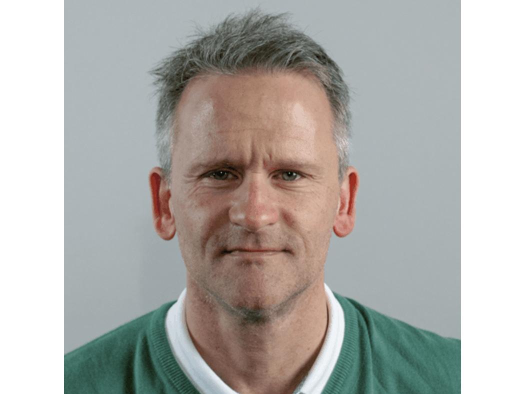 Person wearing a green sweater over a white collared shirt, positioned against a plain light gray background.
