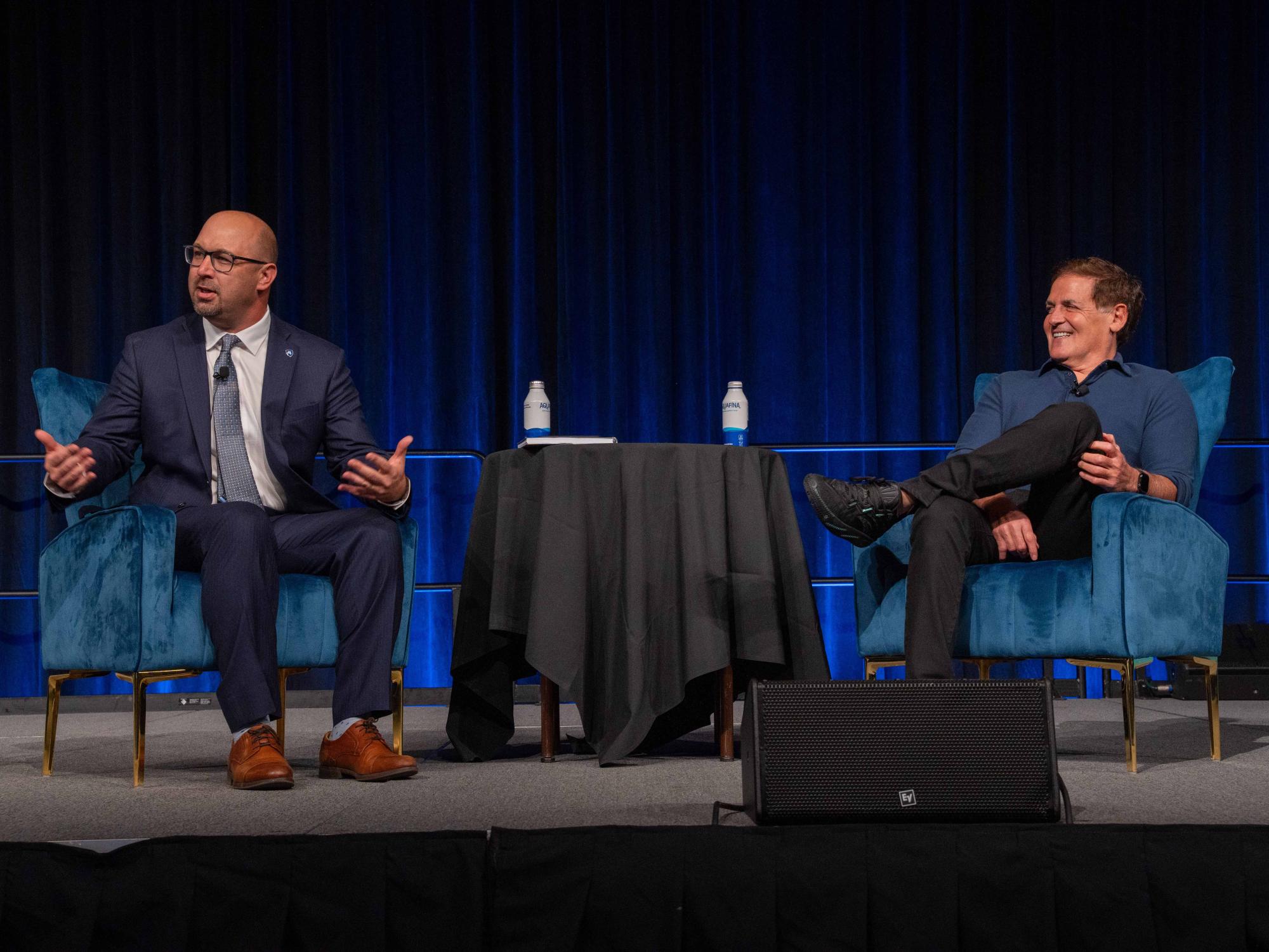 Two speakers seated on stage in blue armchairs with a small round table between them.