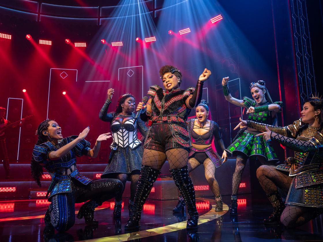 A Black woman leads a diverse cast of women singing into a microphone.