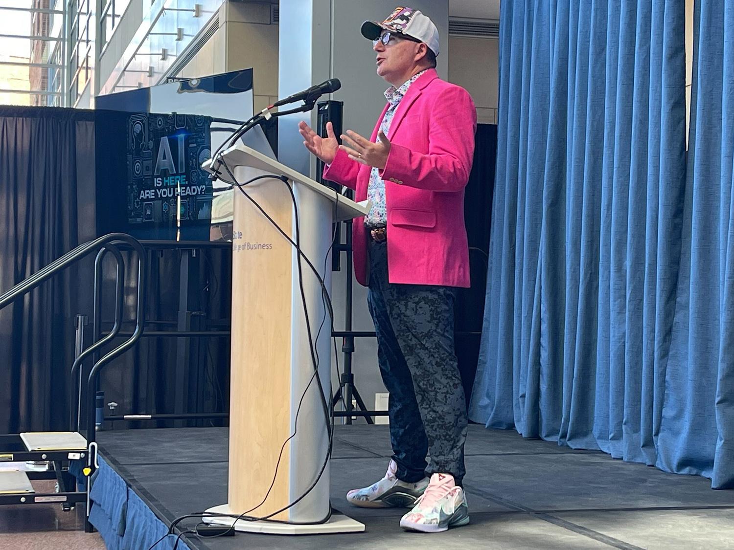 Dan Ives, wearing a hat, glasses, pink blazer and sneakers, speaks into a microphone from behind a lectern