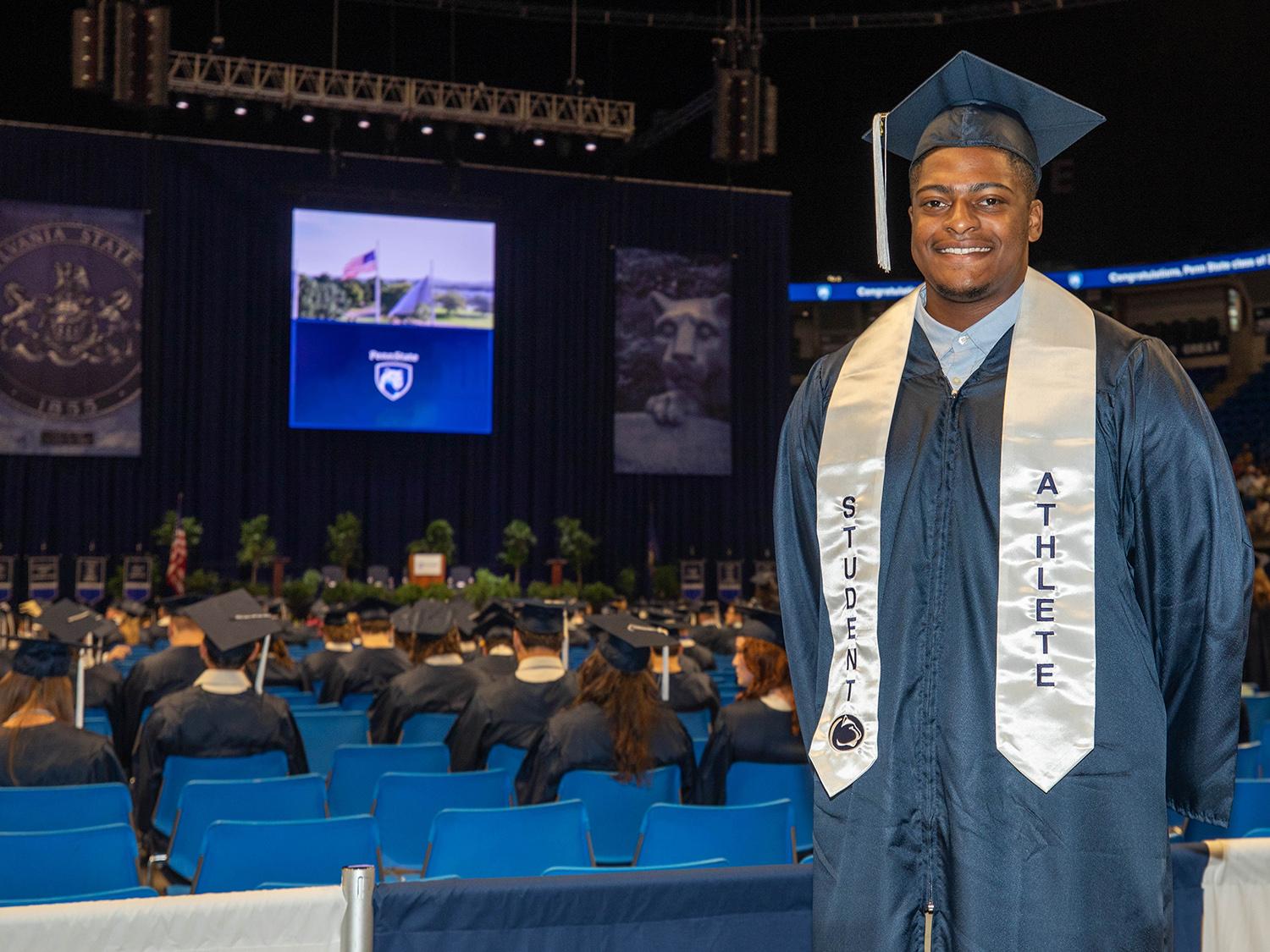 Gabe Nwosu stands in a blue cap and gown with a white sash that reads Student Athlete prior to one of the two Penn State Smeal College of Business commencement ceremonies he has attended. 