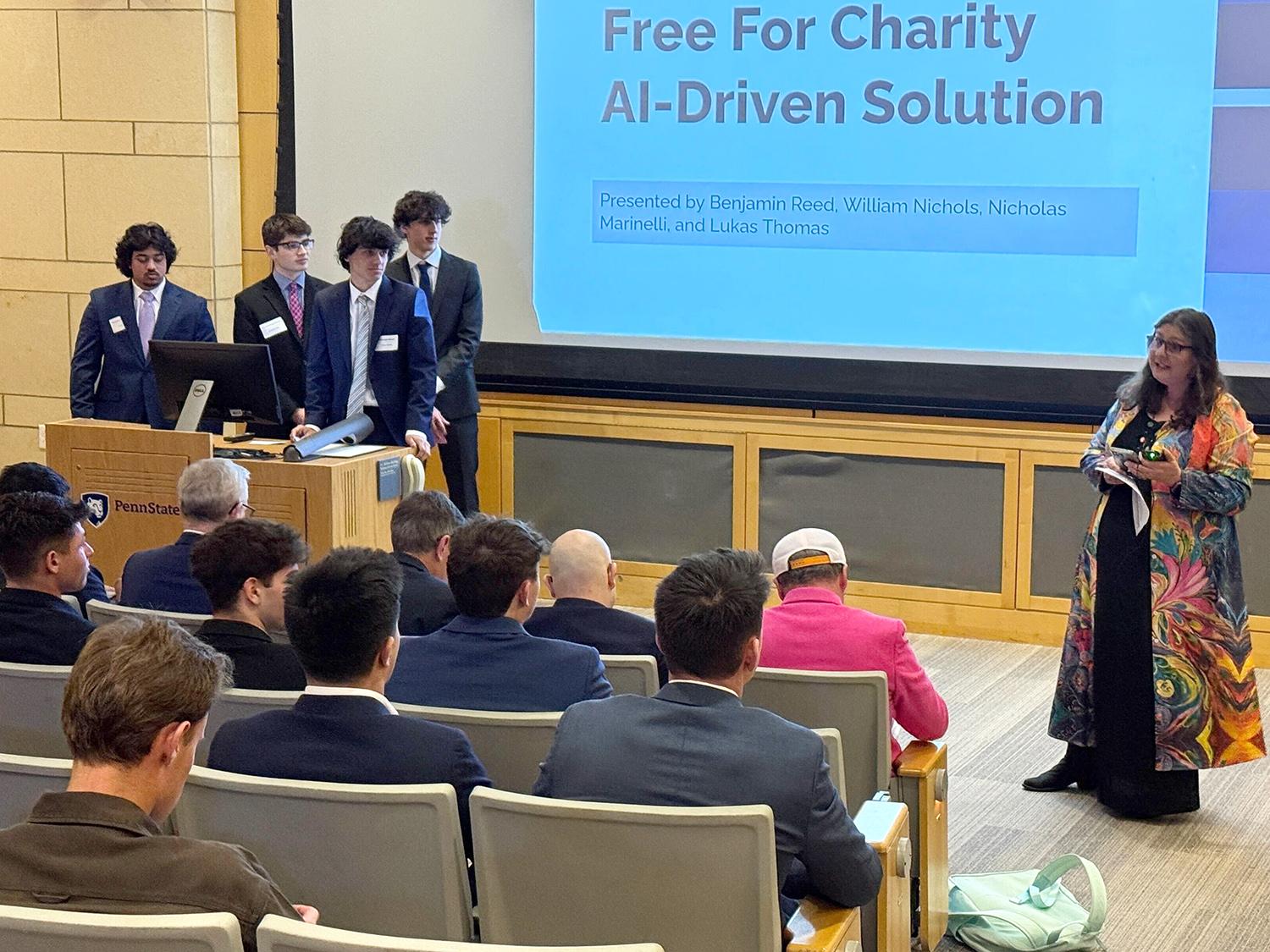 Four male students dressed in suits stand behind a lectern in the Struthers Auditorium in the Penn State Business Building as a woman to their left addressed the assembled crowd.