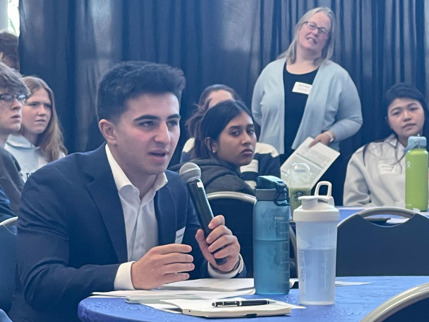 A student holding a microphone and sitting at a table asks a question amid a crowd of students in the Business Building atrium.