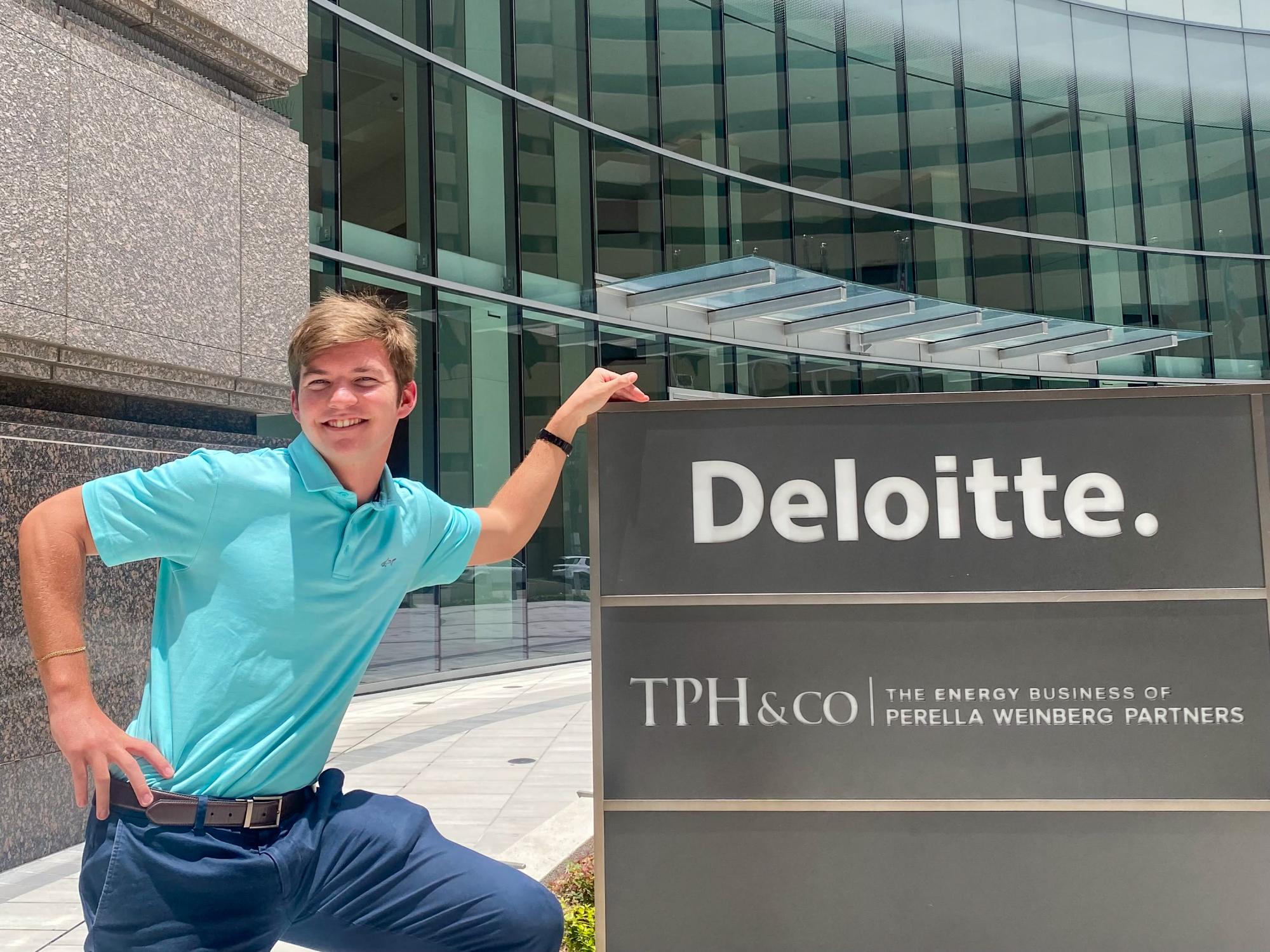 Morgan Overman wears an aqua shirt and navy pants while standing next to a TPH&Co. and Deloitte sign. 
